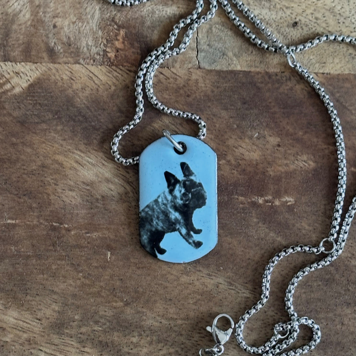 Ona Chan Jewelry | pet tag | dog tag custom enamel dog tag necklace with a black and white French Bulldog on a pastel light blue background, displayed on a rustic wooden surface.