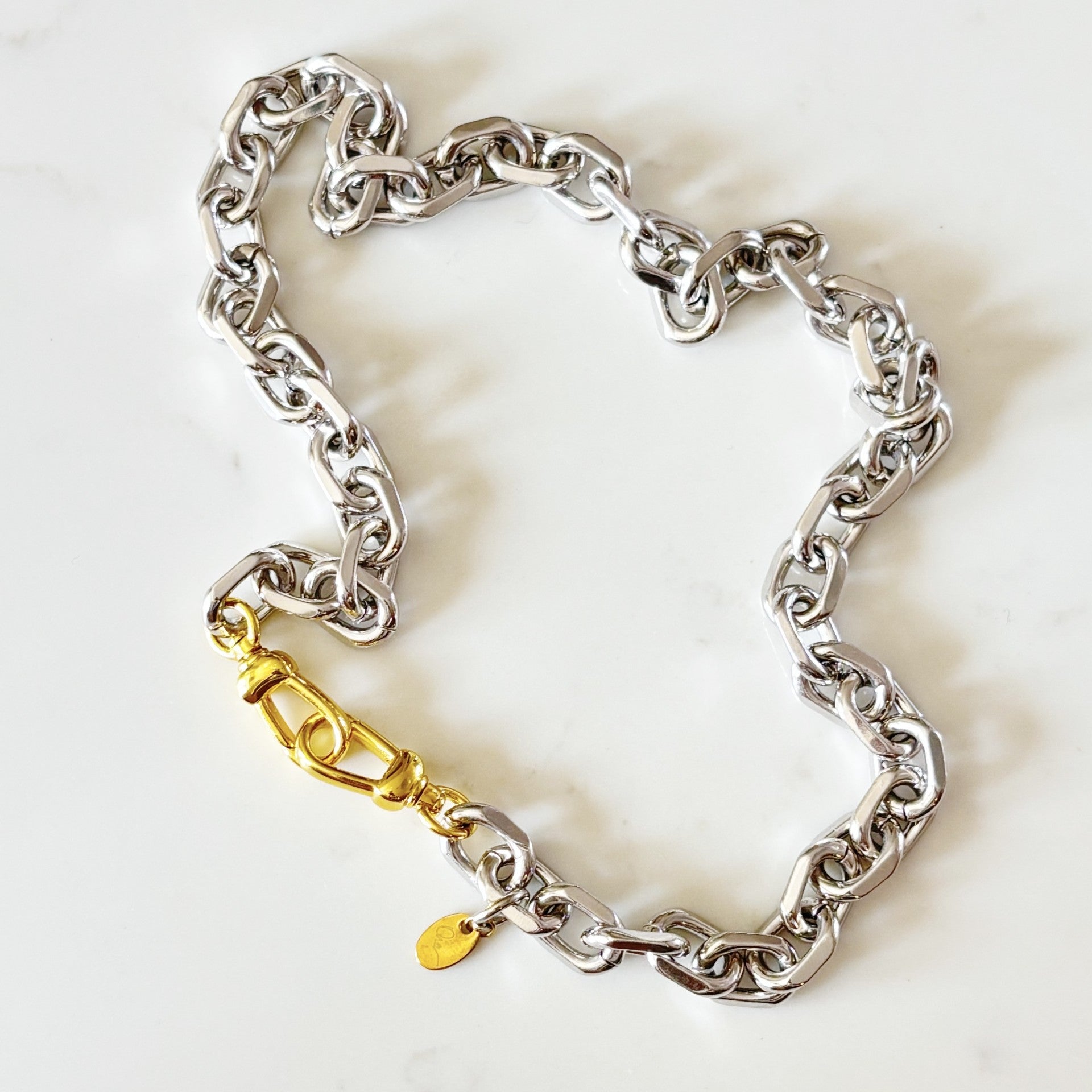 Riley chunky silver-toned chain bracelet with a 18K plated push clasp and a small gold tag, displayed on a light marble background.
