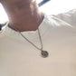 A close-up of a person wearing a white short-sleeve crewneck t-shirt and a wearing H initial stamp charm in fine silver with a steel box chain necklace 
