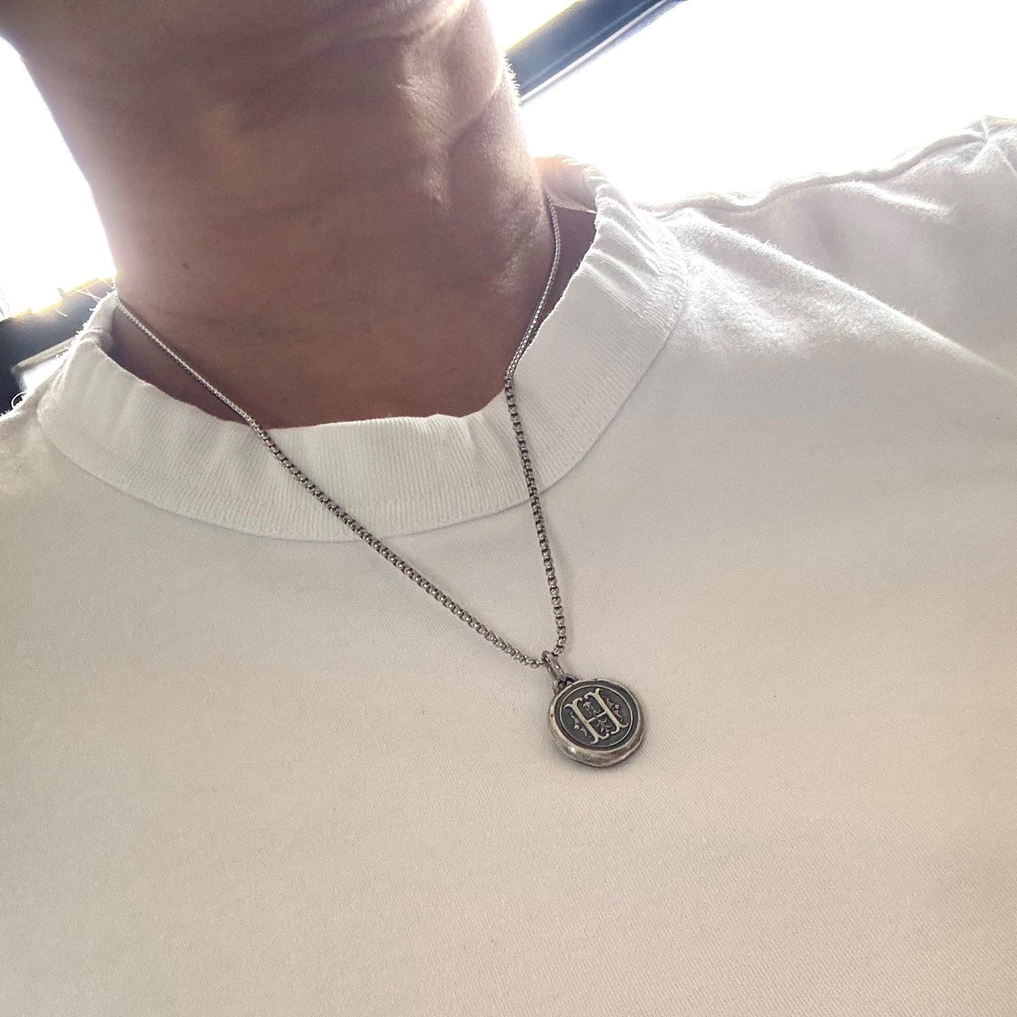 A close-up of a person wearing a white short-sleeve crewneck t-shirt and a wearing H initial stamp charm in fine silver with a steel box chain necklace 
