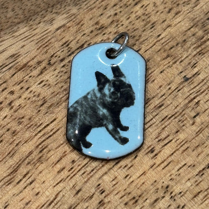 A pastel blue vitreous enamel pet tag | dog tag pendant featuring a black and white decal, resting on a textured wooden surface.