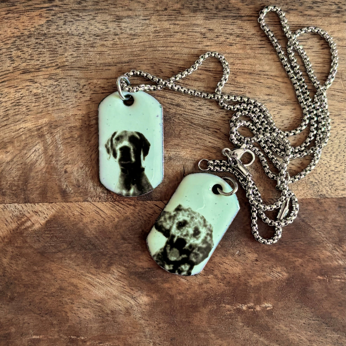 Two Ona Chan Jewelry dog tag necklaces with enamel dog custom portraits with steel box chains, displayed on a rustic wooden surface. featuring custom black and white photo of dogs in a light green background with 