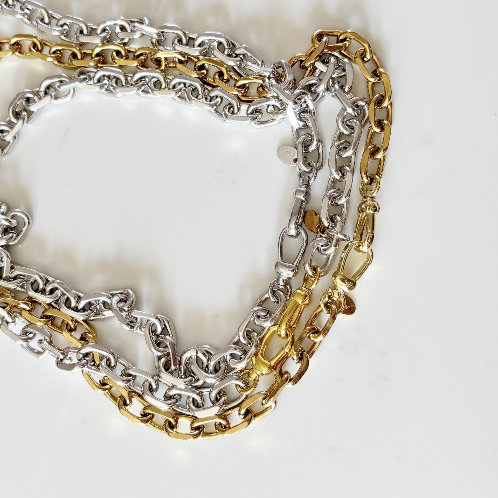 A close-up of three Rileyn interwoven chain necklaces, one silver-toned and one gold-toned, one Silver-tone with silver claps arranged on a light off-white background.