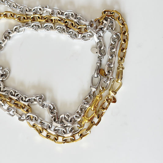 A close-up of three Rileyn interwoven chain necklaces, one silver-toned and one gold-toned, one Silver-tone with silver claps arranged on a light off-white background.
