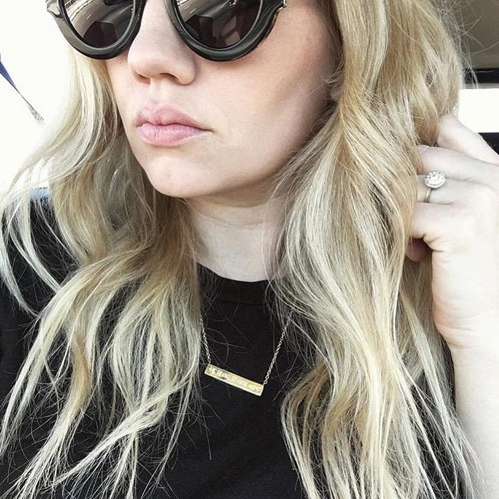 Blond woman wearing black sunglasses and shirt wearing gold Mantra Engraved Horizontal Necklace